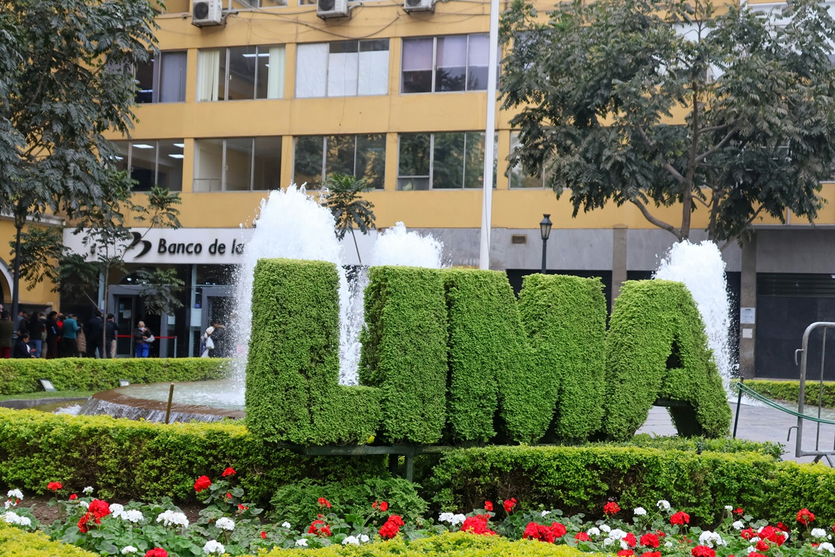 Topiaria em formato das letras “LIMA” feita de folhagem verde diante de fonte jorrando água, com edifício e árvores ao fundo
